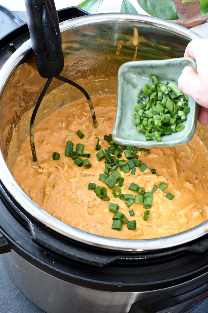 Shredded instant pot buffalo chicken dip with a hand pouring in sliced green onion.