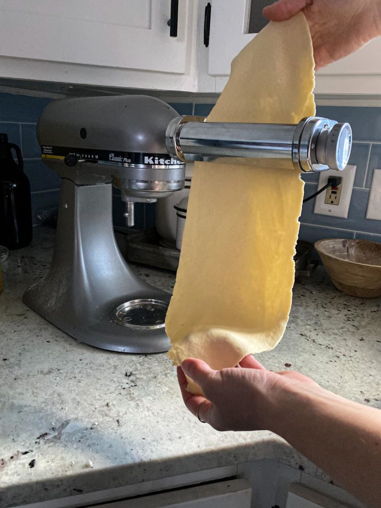 A kitchen aid stand mixer on a white counter with a flat sheet of dough being put through it