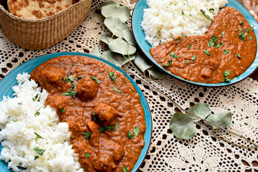 Two blue plates willed with orange veggie tikka masala with white rice and naan in the image.  The plates are on lace