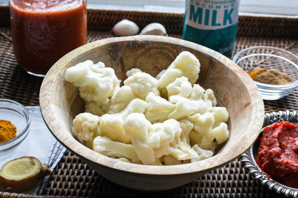 A wooden bowl of frozen cauliflower, with small bowls of spices and tomato paste around it.  There is a can of coconut milk in the background, and it is all on a wicker serving tray
