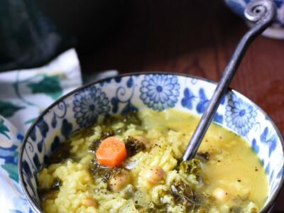 a blue and white flowered bowl filled with greek lemon and rice soup, with carrots, kale and chickpeas visible in the soup. There is another bowl in the background, and they are on a dark wooden surface