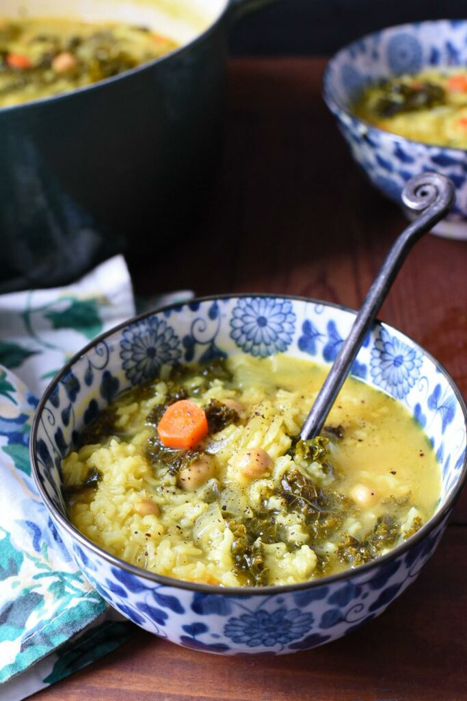 a blue and white flowered bowl filled with greek lemon and rice soup, with carrots, kale and chickpeas visible in the soup.  There is another bowl in the background, and they are on a dark wooden surface