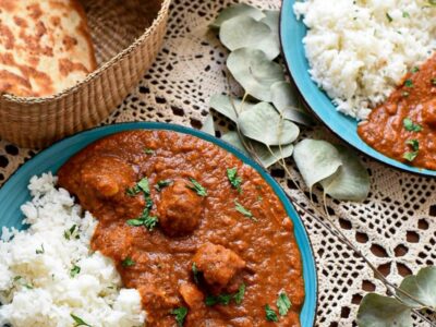 Two blue plates filled with bright orange veggie tikka masala and basmati rice. There is naan in the background in a wicker basket, and lace under the plates