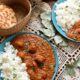 Two blue plates filled with bright orange veggie tikka masala and basmati rice. There is naan in the background in a wicker basket, and lace under the plates