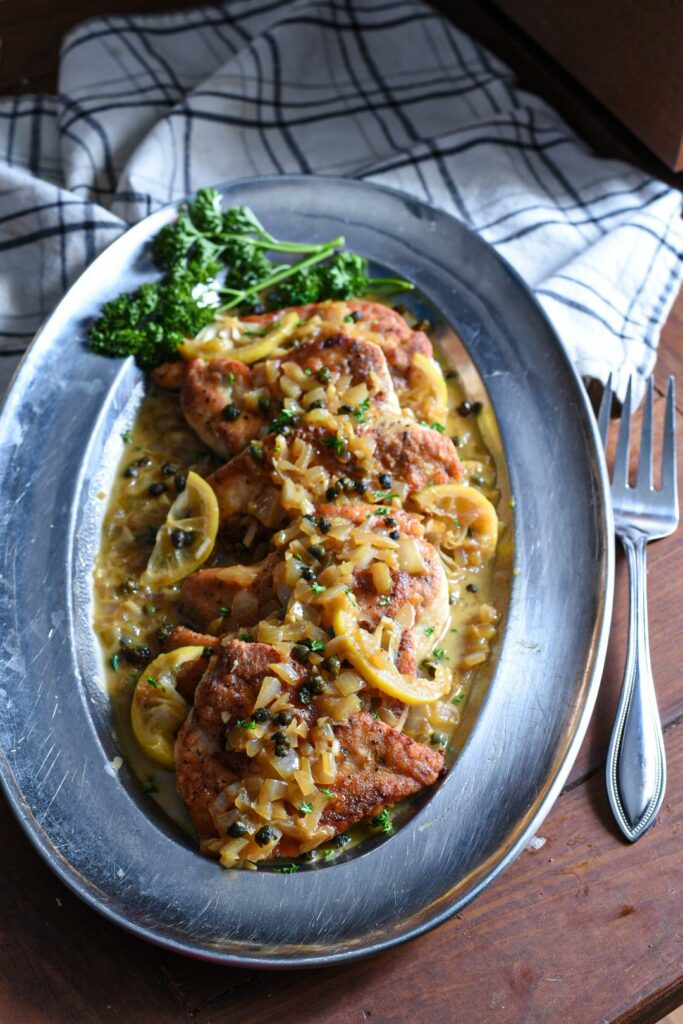 Sliced chicken breasts covered in a chicken piccata sauce, capers, parsley, lemon slices and onion on a sliver serving dish.