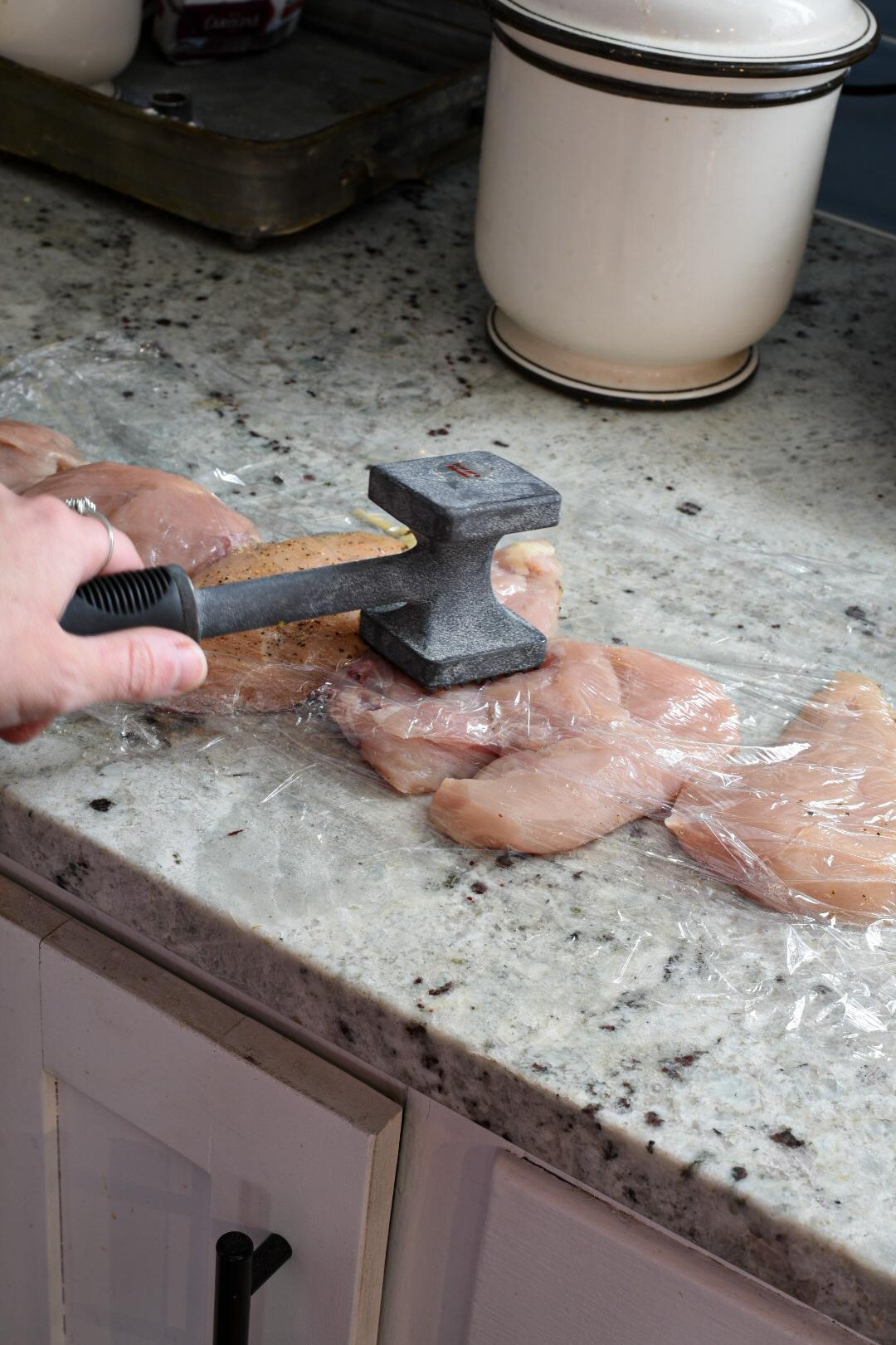 A hand holding a poultry hammer pounding chicken breasts in between two pieces of plastic wrap