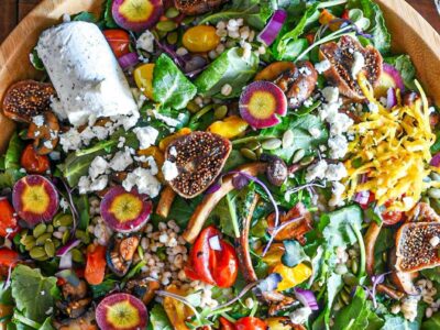 A large wooden serving bowl filled with mushroom and kale winter salad, topped with figs and goat cheese