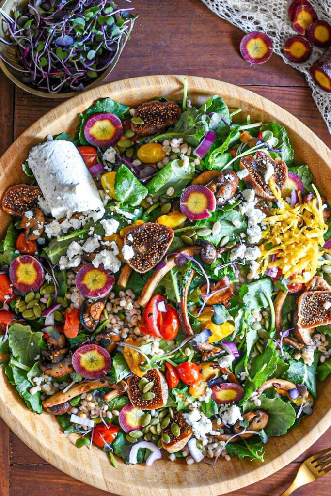 A large wooden serving bowl filled with mushroom and kale winter salad, topped with figs, barley and goat cheese