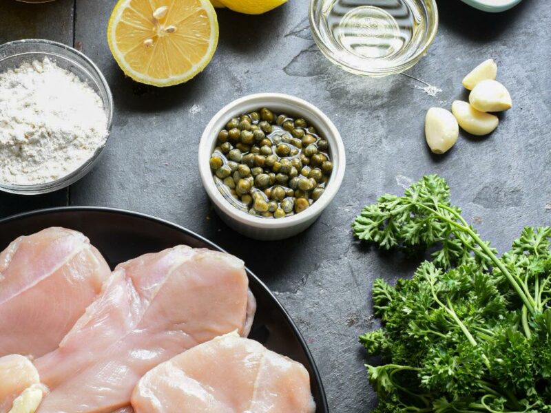 easy chicken piccata recipe An overhead shot of the ingredients needed to make easy chicken piccata including capers chicken breasts and lemons.