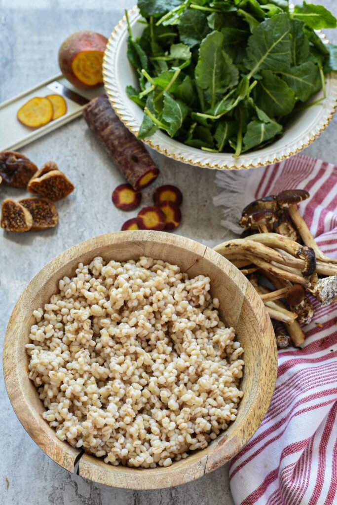 A wooden bowl filled with barley, with baby kale and sliced purple carrot in the background