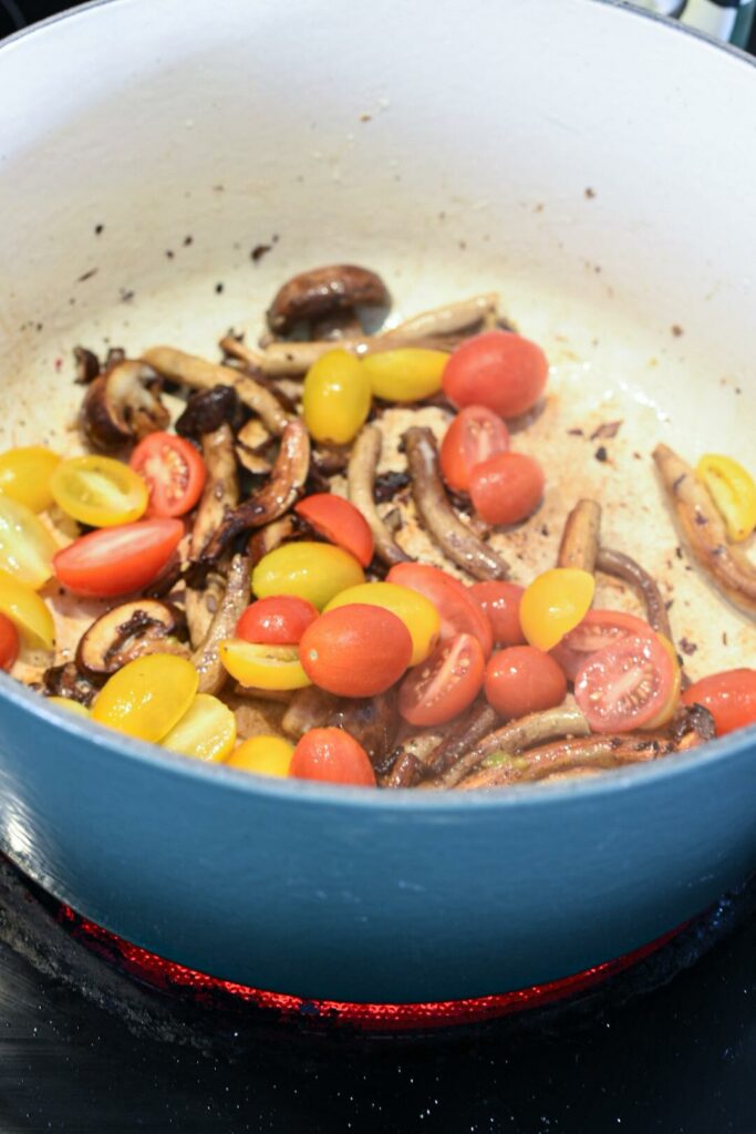 A blue dutch oven on a stove top filled with yellow and red cherry tomatoes and shiitake mushrooms