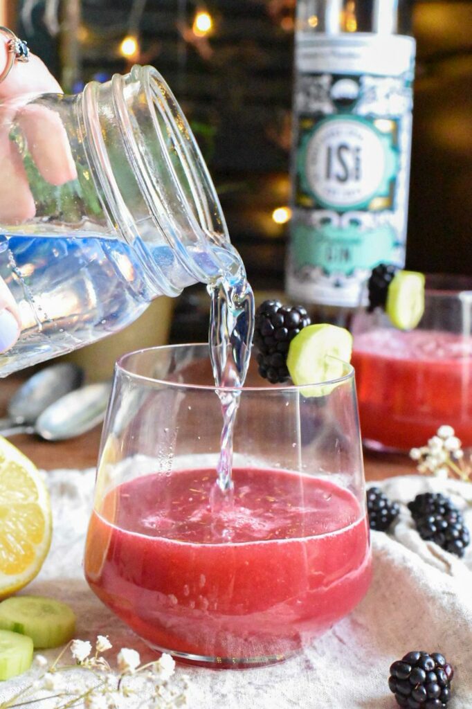 A hand pouring tonic into a bright red cucumber blackberry gin cocktail.