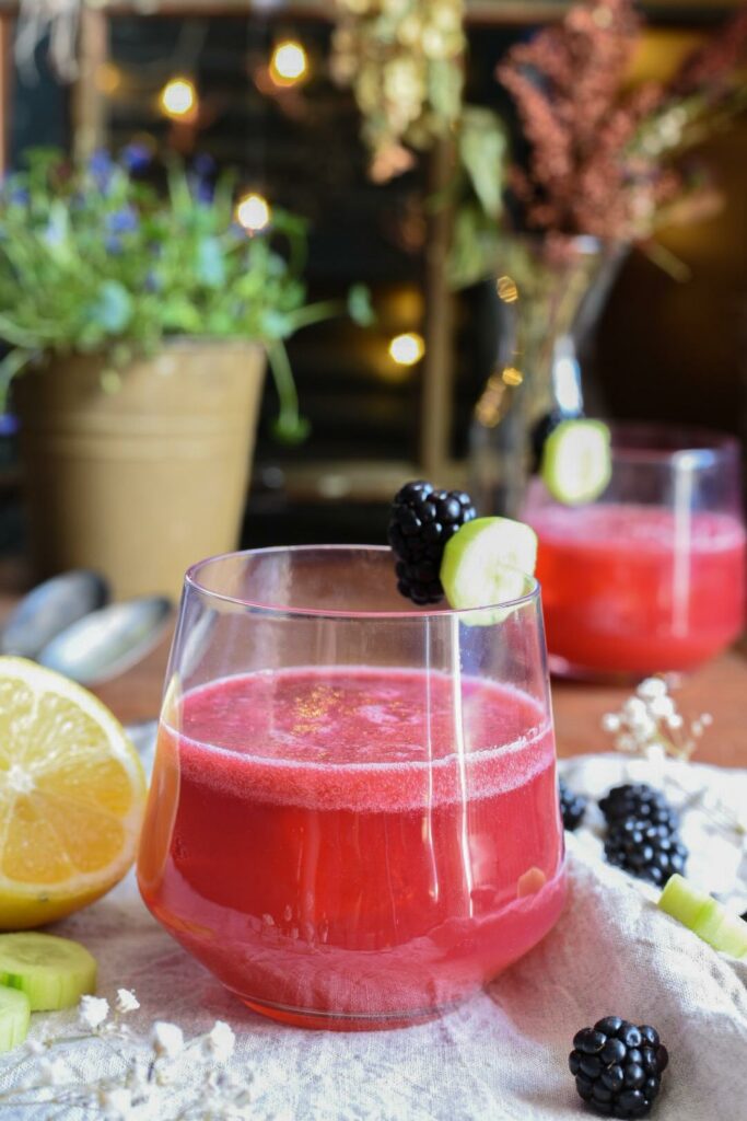 A cucumber blackberry gin cocktail in a glass surrounded by sliced cucumbers, blackberries and a lemon slice.