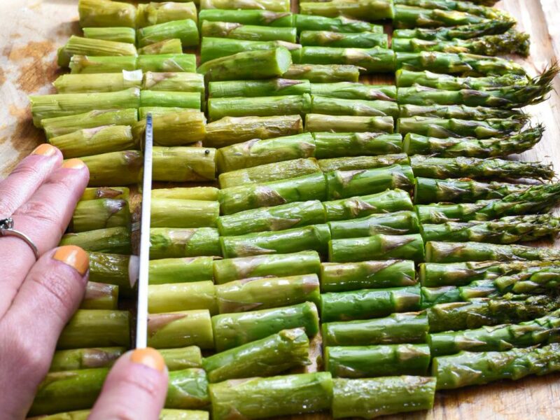 A hand holding a knife cutting asparagus into small pieces on a wooden cutting board