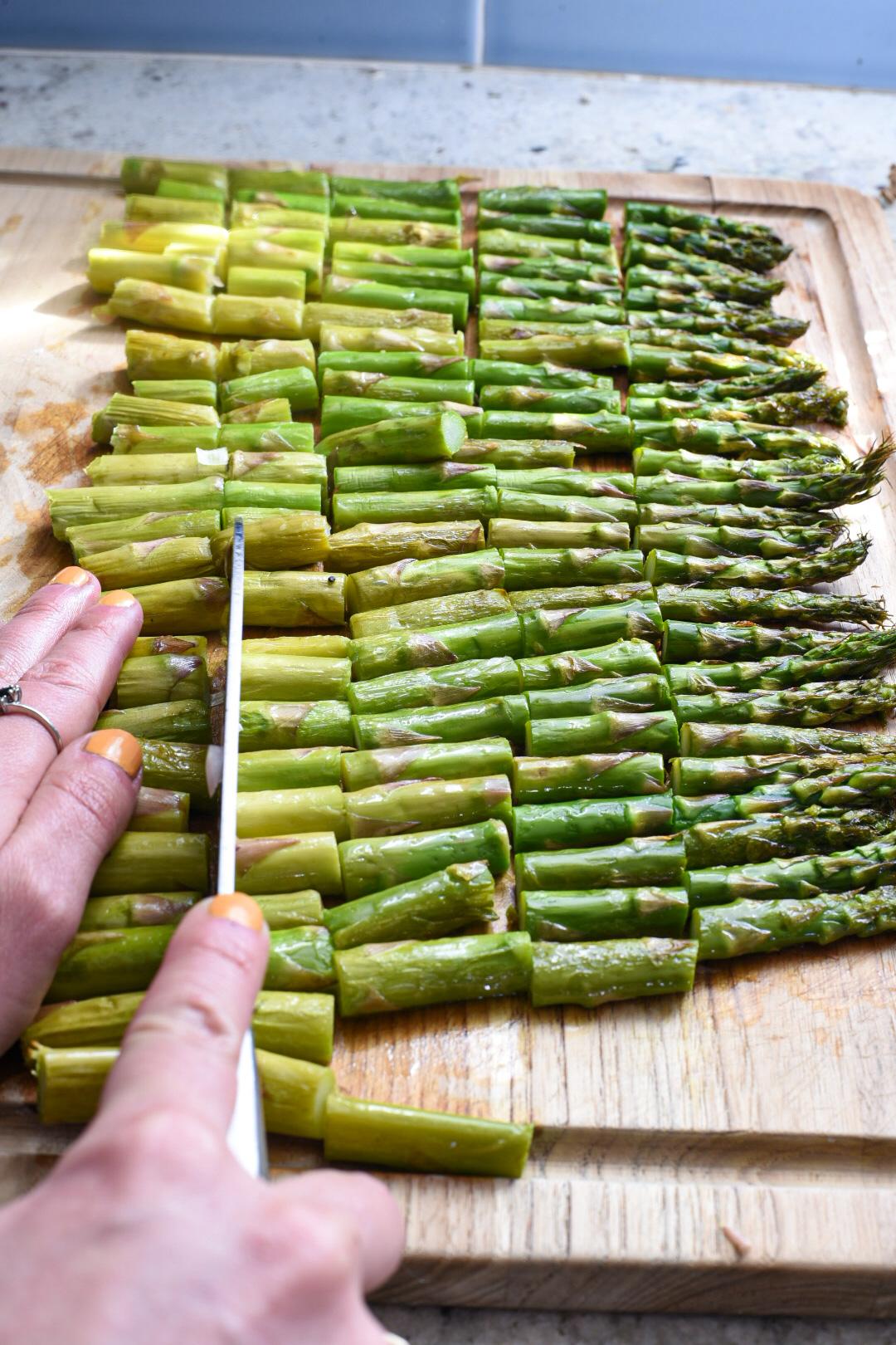 A hand holding a knife cutting asparagus into small pieces on a wooden cutting board