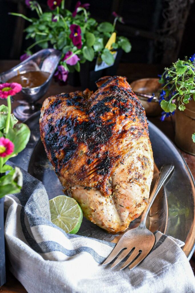 A baked jerk turkey breast on a silver platter with gravy and flowers in the background.