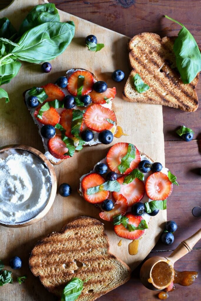 A wooden cutting board with two pieces of blueberry strawberry ricotta toast on it topped with fresh basil, with a small bowl of ricotta cheese next to it