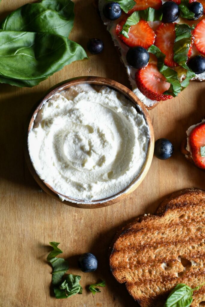A small wooden bowl of whipped ricotta cheese on a wooden cutting board with ricotta toast topped with strawberries, blueberries and basil around it