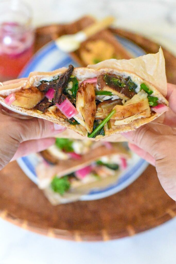 Two hands holding a stuffed chicken pita sandwich with chicken, pickled red onion, shiitakes, spinach and sauce visible.