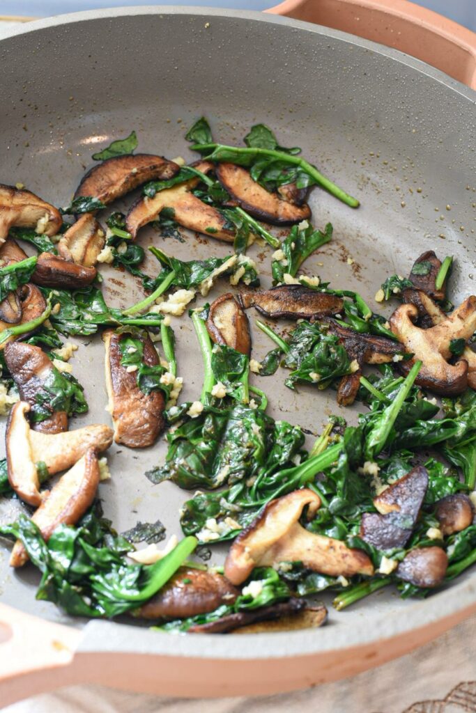 A non-stick pan with cooking golden brown shiitake mushrooms, spinach and garlic