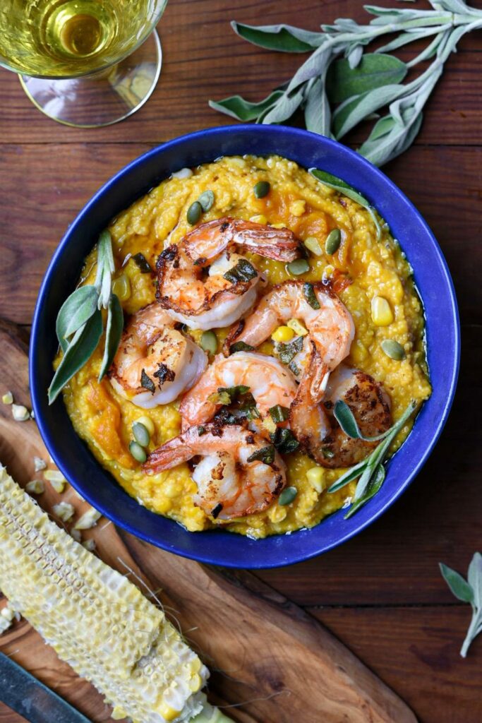 A close up image of brown butter pumpkin polenta in a blue bowl topped with sage ginger shrimp, pumpkin seeds and fresh sage leaves on a wooden table, and a glass of wine in the shot