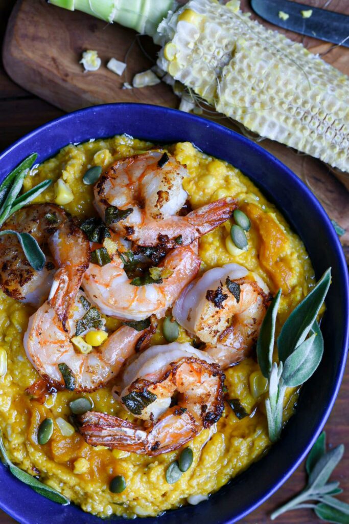 A close up image of brown butter pumpkin polenta in a blue bowl topped with sage ginger shrimp, pumpkin seeds and fresh sage leaves over a wooden table.