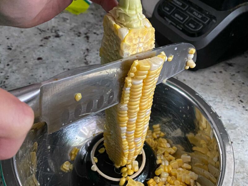 A hand holding the stem of a cob of corn, with the corn positioned on a small bowl inside of a big bowl. There is a knife cutting through the kernels of the corn as they fall into the bowl.