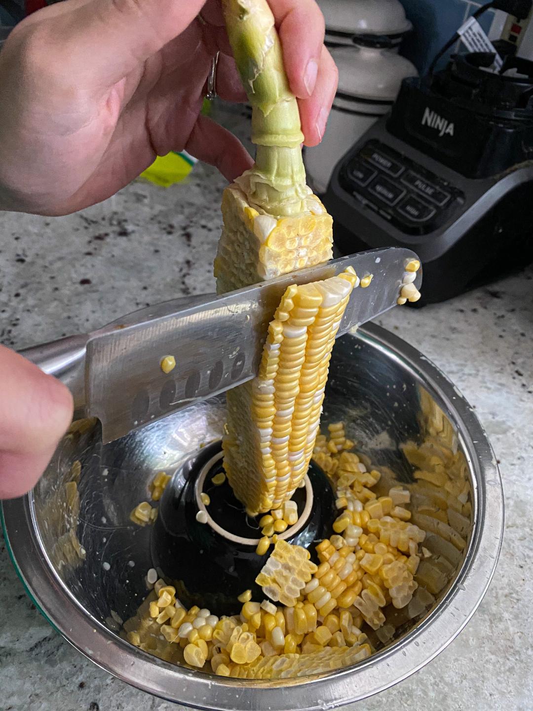 Chopping corn on the cob A hand holding the stem of a cob of corn, with the corn positioned on a small bowl inside of a big bowl. There is a knife cutting through the kernels of the corn as they fall into the bowl.