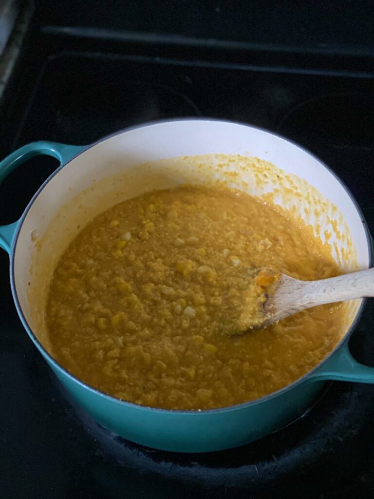 A blue dutch oven filled with cheesy polenta and corn with a wooden spoon in the pot