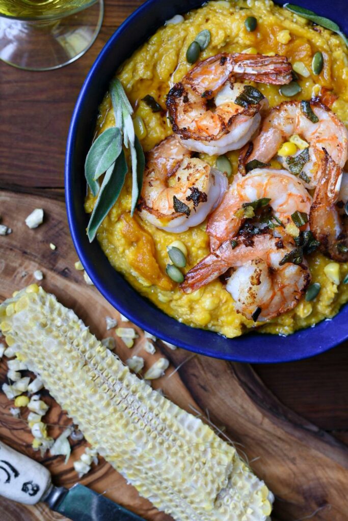 An image of brown butter pumpkin polenta in a blue bowl topped with sage ginger shrimp, pumpkin seeds and fresh sage leaves over a wooden table with a corn cob next to the bowl