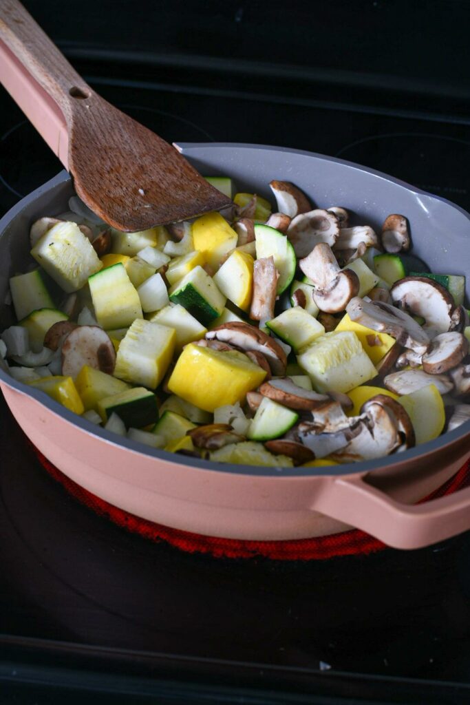 Sauteed zucchini and squash and mushroom in a pink sautee pan with a wooden spoon.