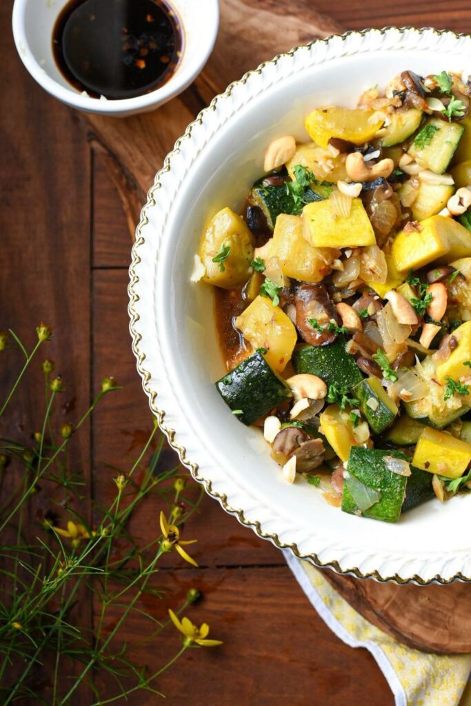 Half of a bowl of zucchini and squash recipe with chopped peanuts and parsley on top and yellow flowers on the table.
