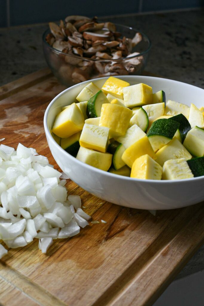 A white bowl filled with zucchini and summer squash, with chopped onion and mushrooms on a wooden cutting board.