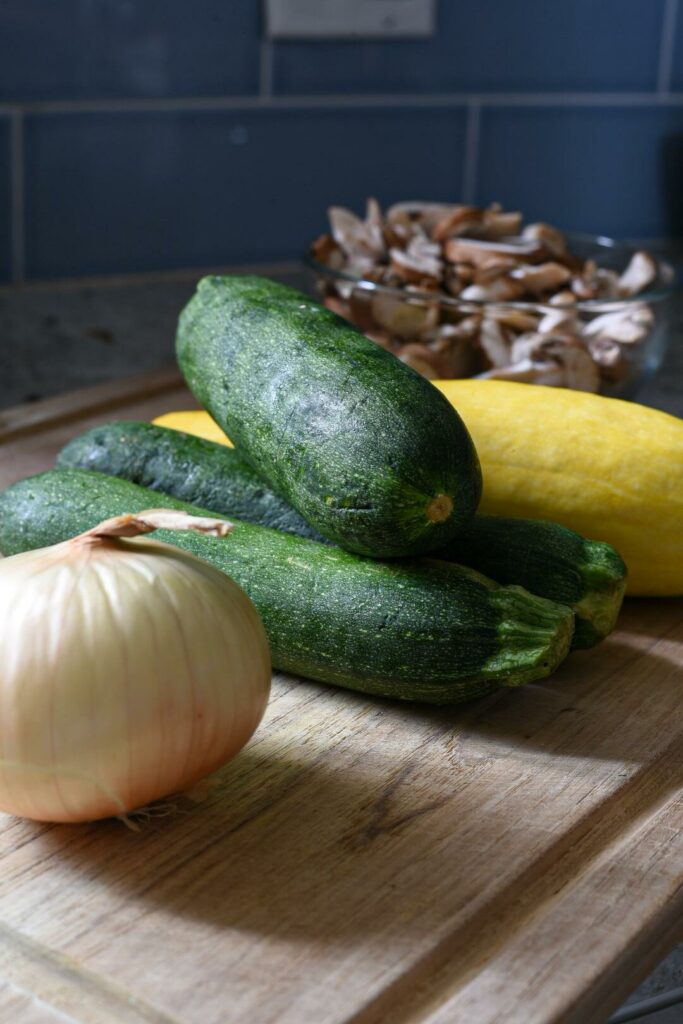 Three whole zucchinis in natural light on a wooden cutting board with onion and yellow squash.