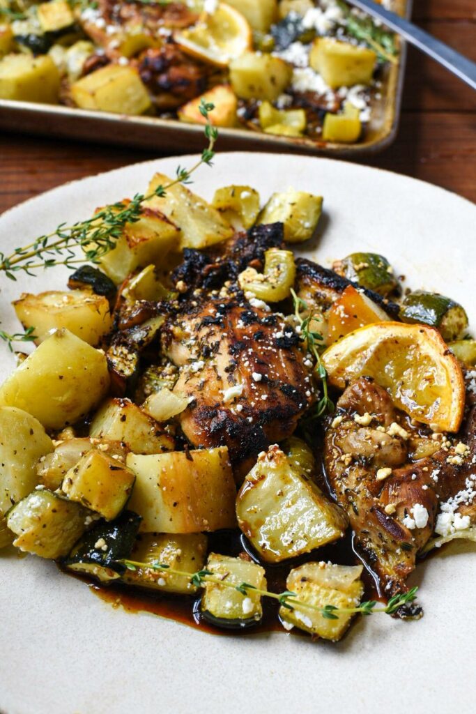 A white plate filled with lemon garlic greek chicken and oven roasted vegetables, roasted lemon rinds, sprinkled feta cheese and thyme.
