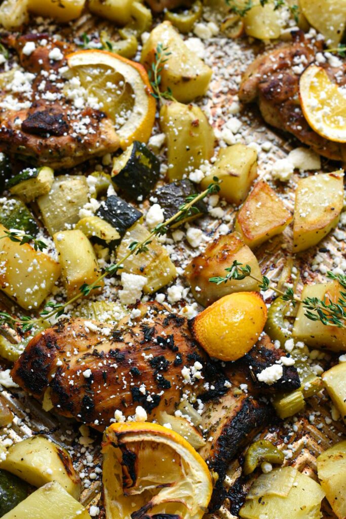 Close up image of sheet pan greek chicken with roasted potatoes, vegetables and bright yellow lemon rinds with charred edges.