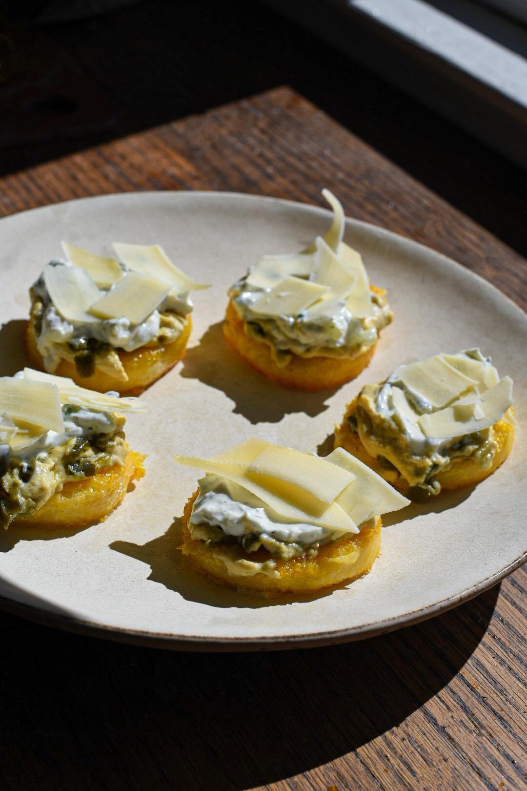 crispy polenta cakes topped with hummus, tzatziki and Parmesan cheese shreds, on a brown wooden table
