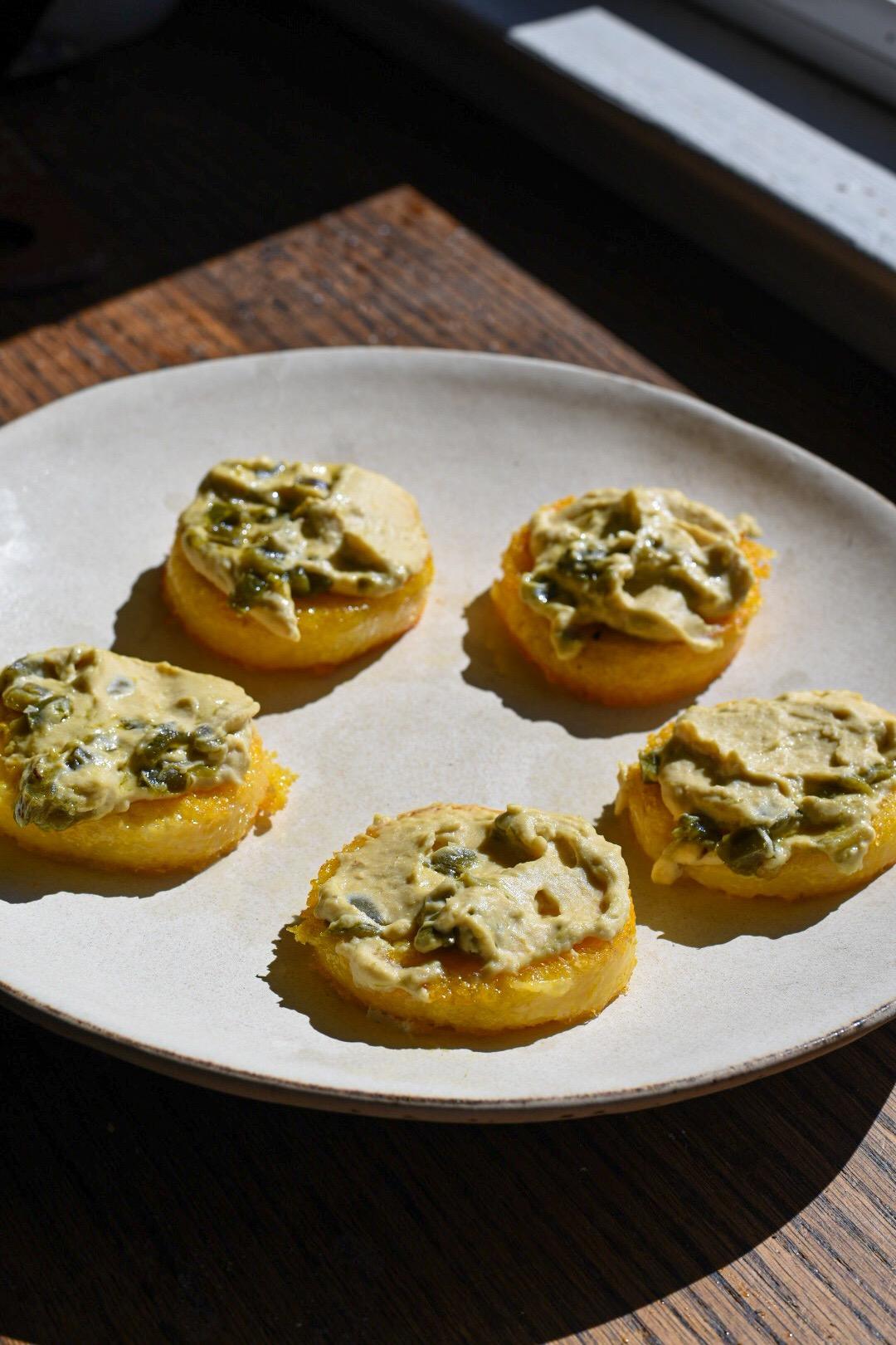 crispy polenta cakes topped with hummus on a white plate in the sun