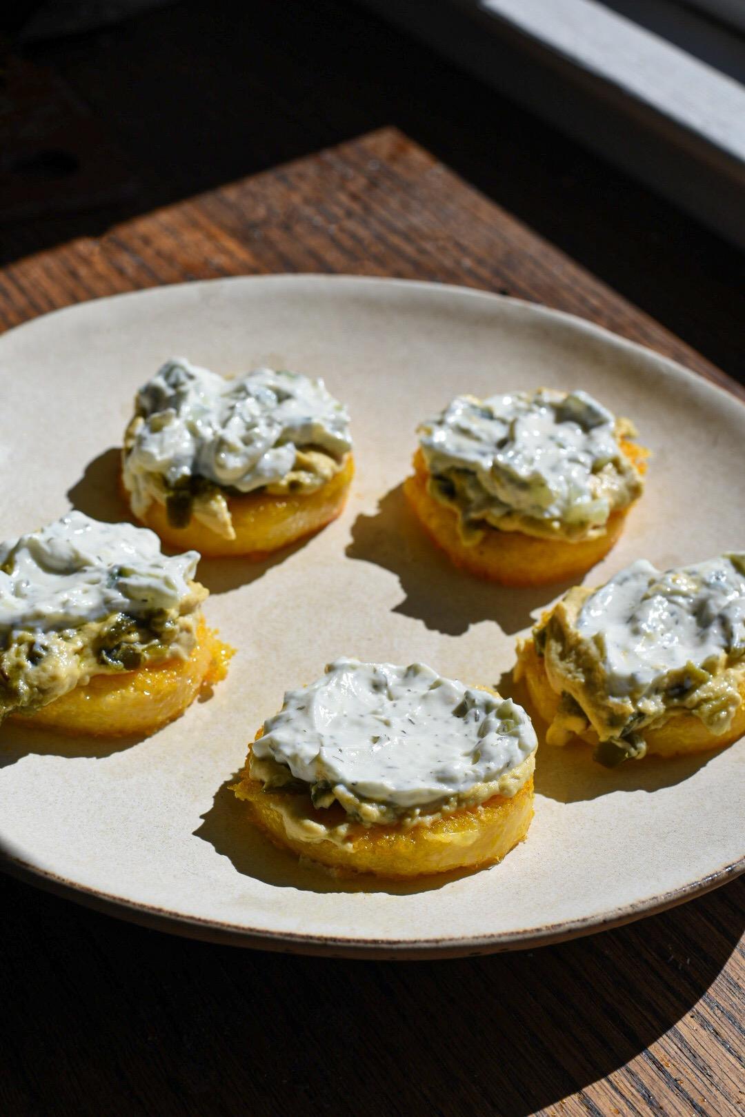 polenta rounds topped with hummus and tzatziki on a white plate on a wooden table