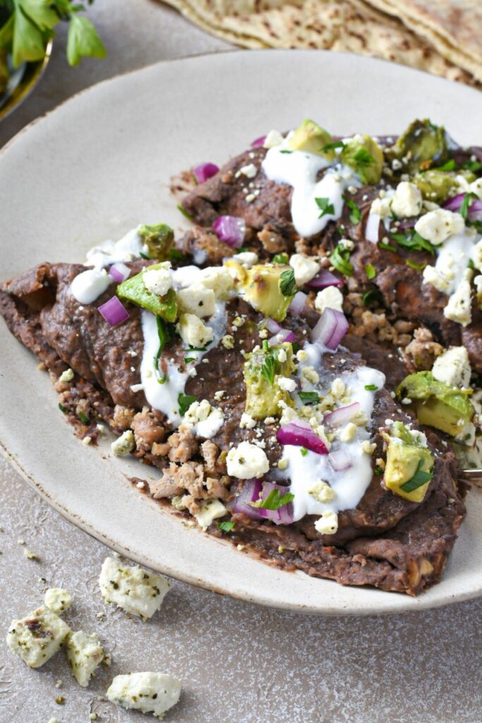 two enfrijoladas on a white plate covered in a black bean sauce, with red onion, avocado and feta cheese sprinkled over the top.