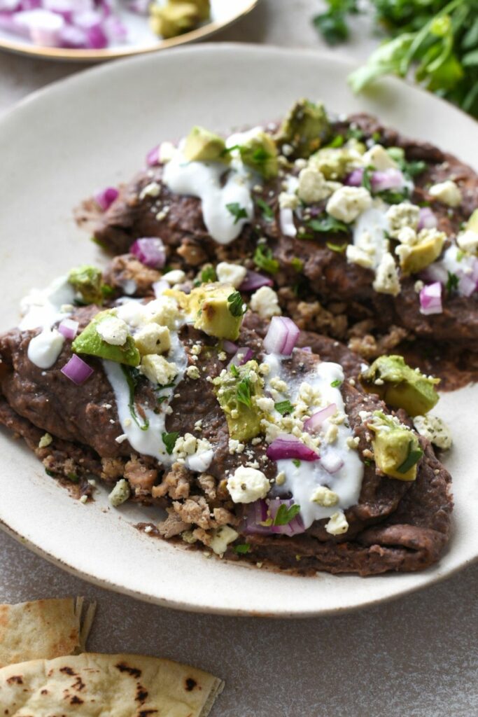 A white plate with two enfrijoladas pita pockets filled with ground turkey and topped with a black bean sauce, red onion, feta, avocado and cilantro. There are two pita pockets in the corner of the image