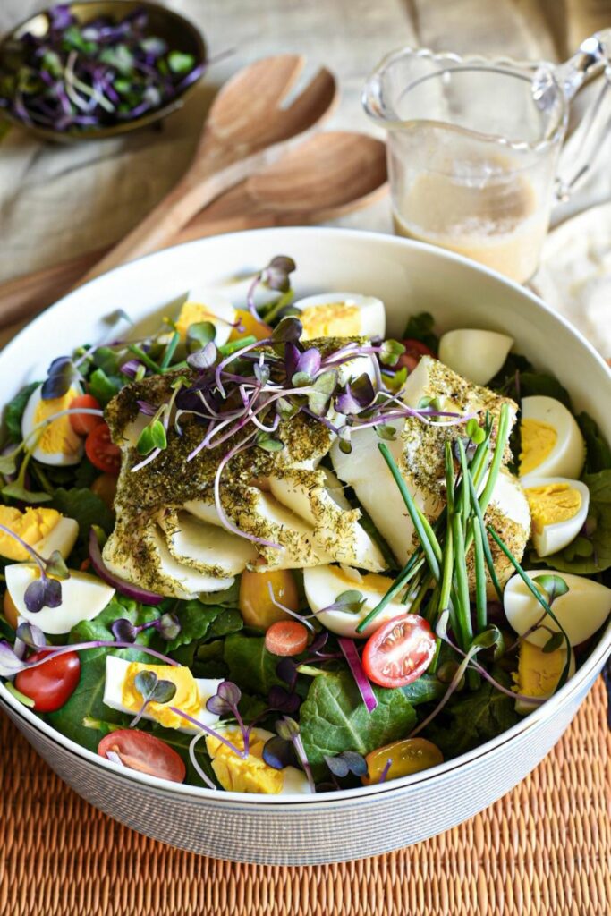 A large bowl of salad with a piece of cooked dill cod fish, with chopped hard boiled eggs, baby kale, tomatoes and microgreens in the salad. There is a homemade caesar salad dressing in the background
