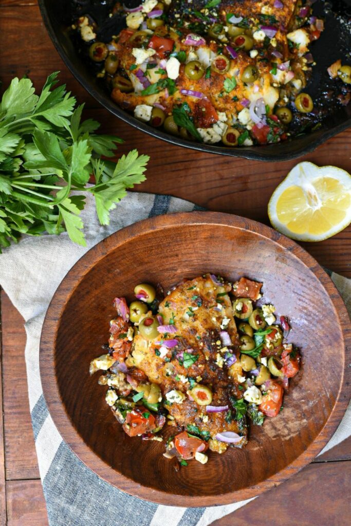 A wooden bowl filled with mediterranean baked cod recipe, with a skillet of the same recipe in the background