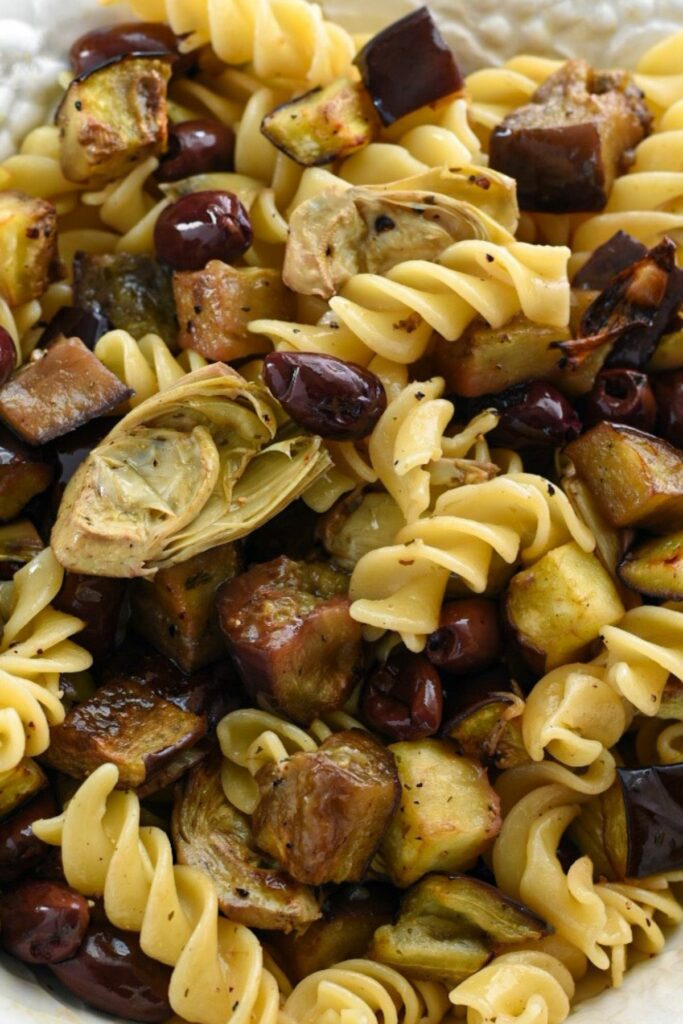 A close up shot of cooked fusilli pasta tossed with roasted eggplant, artichoke hearts and kalamata olives