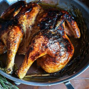 A cooked spatchcock chicken in a large stainless steel skillet with crispy brown skin and rosemary under it.