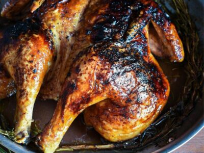 A cooked spatchcock chicken in a large stainless steel skillet with crispy brown skin and rosemary under it.