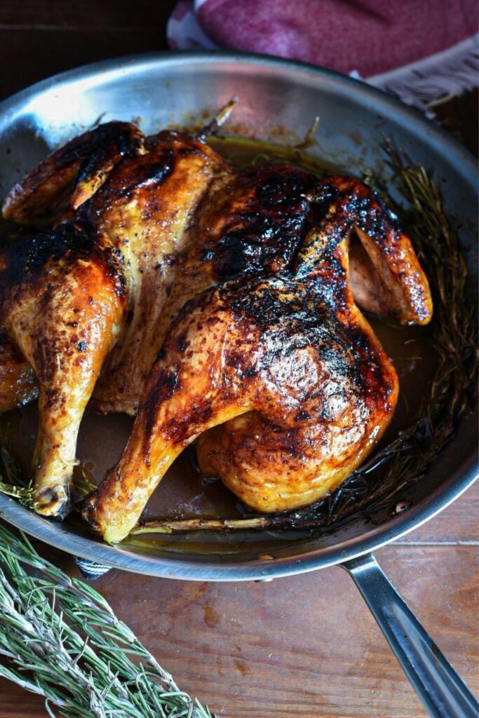 A cooked spatchcock chicken in a large stainless steel skillet with crispy brown skin and rosemary under it.