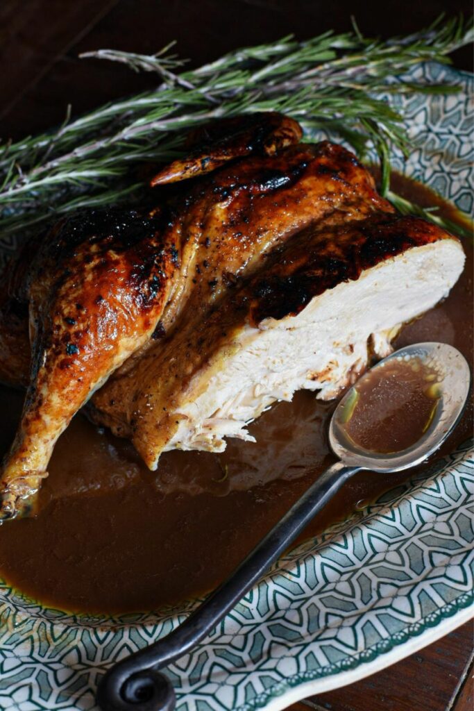 A whole roasted pomegranate chicken cut in half so you can see the cooked white meat inside on a swerving platter with pomegranate gravy and rosemary sprigs