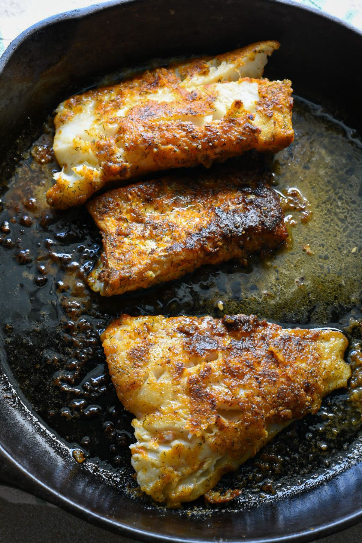 A large cast iron skillet filled with three golden pan seared cod fillets, and a butter olive oil sauce