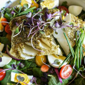 A close up shot of a large serving bowl filled with baby kale salad, with chopped hard boiled eggs, cherry tomatoes, chives, microgreens and baked cod fish covered in dill over the top